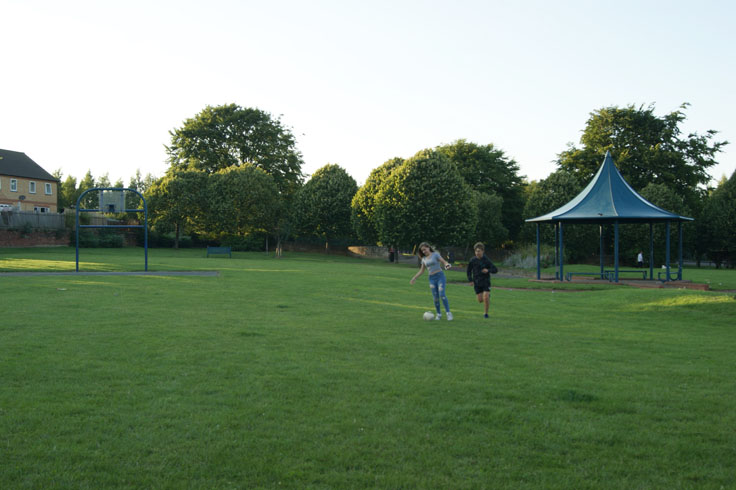 Young people playing football in a park, with a bandstand on their right.
